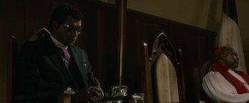 Movie still from “Come Sunday” (2018), directed by Joshua Marston – A man sitting at a table in front of a wooden chair; Medium shot, Low angle