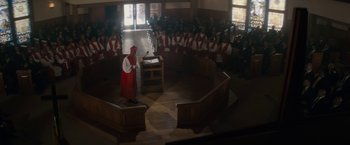 Movie still from “Come Sunday” (2018), directed by Joshua Marston – A man in a red robe stands at a podium in front of a group of people; Extreme Wide shot, High angle