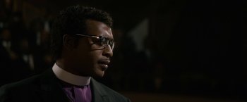 Movie still from “Come Sunday” (2018), directed by Joshua Marston – A person wearing glasses and a suit; Close Up shot, Over the shoulder angle