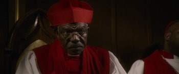 Movie still from “Come Sunday” (2018), directed by Joshua Marston – An older man wearing a red hat and a red robe; Close Up shot, Low angle