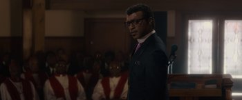 Movie still from “Come Sunday” (2018), directed by Joshua Marston – A man standing in front of a group of people in a room; Close Up shot, Low angle