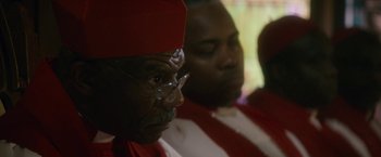 Movie still from “Come Sunday” (2018), directed by Joshua Marston – A man wearing a red hat and glasses; Close Up shot, Over the shoulder angle