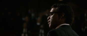 Movie still from “Come Sunday” (2018), directed by Joshua Marston – A person wearing glasses and a suit; Close Up shot, Over the shoulder angle