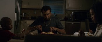 Movie still from “Come Sunday” (2018), directed by Joshua Marston – A man sitting at a table cutting food with a knife and fork; Medium shot, Low angle
