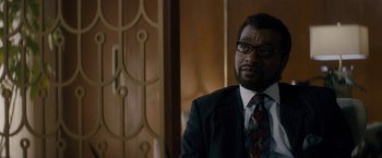 Movie still from “Come Sunday” (2018), directed by Joshua Marston – A man in a suit and tie sitting in front of a door; Close Up shot, Over the shoulder angle