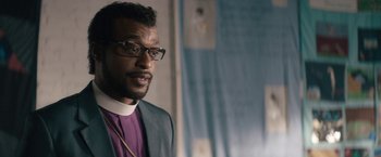 Movie still from “Come Sunday” (2018), directed by Joshua Marston – A person wearing glasses and a suit; Close Up shot, Over the shoulder angle
