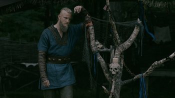 Movie still from “Vikings” (2013), created by Michael Hirst – A man in a viking garb standing next to a tree with skulls hanging from it; Medium shot, Low angle