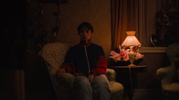 Movie still from “Copenhagen Cowboy” (2022), created by Nicolas Winding Refn – A person sitting in a chair in a living room; Medium shot, High angle