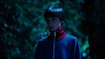 Movie still from “Copenhagen Cowboy” (2022), created by Nicolas Winding Refn – A person standing in the dark with trees in the background; Close Up shot, High angle