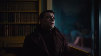 Movie still from “Copenhagen Cowboy” (2022), created by Nicolas Winding Refn – A man in a brown coat sitting in front of a book case; Medium shot, Low angle