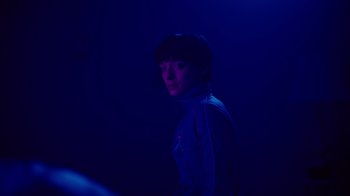 Movie still from “Copenhagen Cowboy” (2022), created by Nicolas Winding Refn – A person standing in the dark wearing a blue jacket; Medium shot, Over the shoulder angle