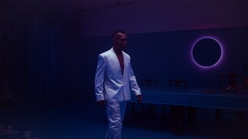 Movie still from “Copenhagen Cowboy” (2022), created by Nicolas Winding Refn – A man in a white suit standing next to a table; Wide shot, Low angle