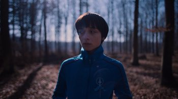 Movie still from “Copenhagen Cowboy” (2022), created by Nicolas Winding Refn – A person in a blue jacket standing in the woods; Close Up shot, Over the shoulder angle