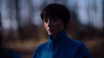 Movie still from “Copenhagen Cowboy” (2022), created by Nicolas Winding Refn – A person wearing a blue jacket standing in a forest; Close Up shot, High angle