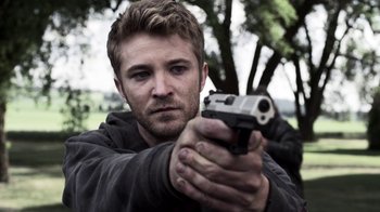 Movie still from “Z Nation” (2014), created by Karl Schaefer – A man holding a gun while standing next to a tree; Close Up shot, Over the shoulder angle