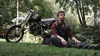 Movie still from “Z Nation” (2014), created by Karl Schaefer – A man sitting on the grass next to a motorcycle; Wide shot, High angle