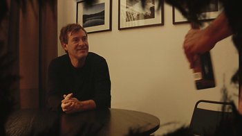 Movie still from “Creep 2” (2017), directed by Patrick Brice – A man sitting at a table in front of a wall of pictures; Medium shot, Over the shoulder angle