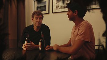Movie still from “Creep 2” (2017), directed by Patrick Brice – Two men sitting at a table with two bottles of beer; Medium shot, Over the shoulder angle