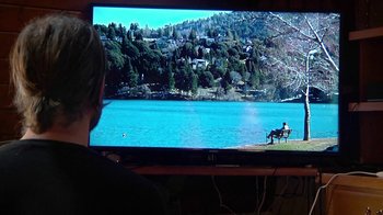 Movie still from “Creep 2” (2017), directed by Patrick Brice – A person sitting on a bench in front of a large screen tv; Wide shot, Over the shoulder angle