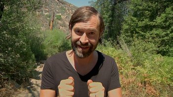 Movie still from “Creep 2” (2017), directed by Patrick Brice – A man with long hair and a beard is giving two thumbs up; Close Up shot, Over the shoulder angle