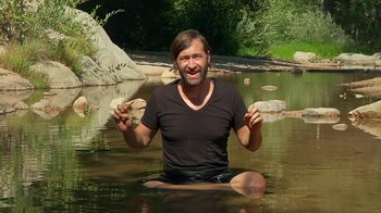 Movie still from “Creep 2” (2017), directed by Patrick Brice – A man sitting in the water holding a piece of food; Medium shot, High angle