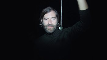 Movie still from “Creep 2” (2017), directed by Patrick Brice – A man with long hair and a goatee is holding a string; Medium shot, Overhead angle