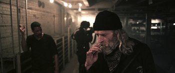 Movie still from “Z Nation” (2014), created by Karl Schaefer – An old man smoking a cigarette while standing next to a wall; Medium shot, Over the shoulder angle