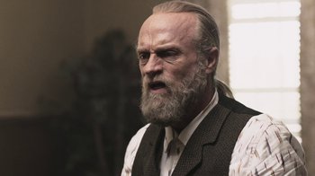Movie still from “Z Nation” (2014), created by Karl Schaefer – An old man with a long beard wearing a vest and a tie; Close Up shot, Low angle