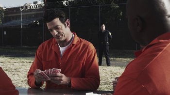 Movie still from “Z Nation” (2014), created by Karl Schaefer – A man in orange shirt playing cards in front of a fence; Medium shot, Over the shoulder angle