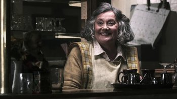 Movie still from “Z Nation” (2014), created by Karl Schaefer – An older woman sitting at a table with a coffee mug; Medium shot, Over the shoulder angle
