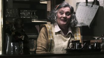 Movie still from “Z Nation” (2014), created by Karl Schaefer – An older woman sitting at a table with a cup of coffee; Medium shot, Over the shoulder angle