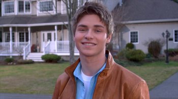 Movie still from “16 Wishes” (2010), directed by Peter DeLuise – A young man smiles for the camera in front of a house; Close Up shot, Over the shoulder angle