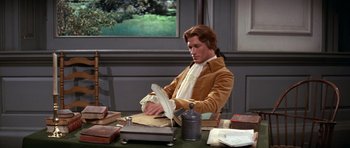 Movie still from “1776” (1972), directed by Peter H. Hunt – A man sitting at a table writing on a paper; Medium shot, High angle