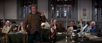 Movie still from “1776” (1972), directed by Peter H. Hunt – A man in a brown and green jacket standing in front of a group of people; Medium shot, Over the shoulder angle