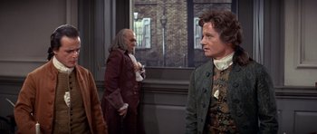 Movie still from “1776” (1972), directed by Peter H. Hunt – A man in a green jacket standing next to another man in a brown jacket; Medium shot, Over the shoulder angle