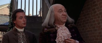 Movie still from “1776” (1972), directed by Peter H. Hunt – A bald man in a wig and a pink jacket; Close Up shot, Over the shoulder angle