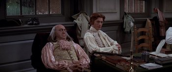 Movie still from “1776” (1972), directed by Peter H. Hunt – A man sitting next to another man in a chair; Medium shot, High angle