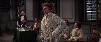 Movie still from “1776” (1972), directed by Peter H. Hunt – A man in a suit and tie standing in front of a group of people; Medium shot, Low angle