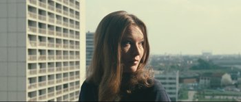 Movie still from “2 or 3 Things I Know About Her” (1967), directed by Jean-Luc Godard – A woman with long brown hair is standing in front of a building; Close Up shot, Low angle