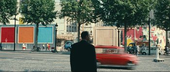 Movie still from “2 or 3 Things I Know About Her” (1967), directed by Jean-Luc Godard – A man standing on a street corner next to a red car; Extreme Wide shot, Over the shoulder angle