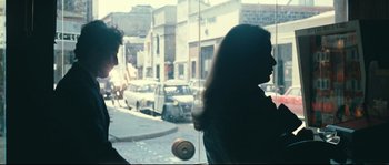 Movie still from “2 or 3 Things I Know About Her” (1967), directed by Jean-Luc Godard – A woman standing in front of a window with a doughnut; Medium shot, Over the shoulder angle