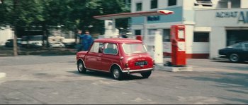 Movie still from “2 or 3 Things I Know About Her” (1967), directed by Jean-Luc Godard – A red car driving down a street next to gas pumps; Extreme Wide shot, High angle