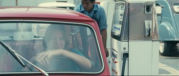Movie still from “2 or 3 Things I Know About Her” (1967), directed by Jean-Luc Godard – A woman sitting in the driver's seat of a red car; Medium shot, Over the shoulder angle