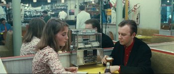 Movie still from “2 or 3 Things I Know About Her” (1967), directed by Jean-Luc Godard – A woman and a man sitting in front of a machine; Medium shot, Over the shoulder angle