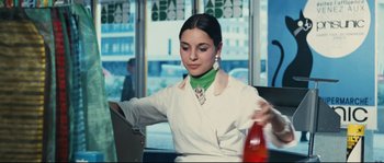 Movie still from “2 or 3 Things I Know About Her” (1967), directed by Jean-Luc Godard – A woman sitting at a table with a bottle of soda; Medium shot, Low angle
