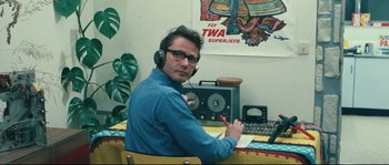 Movie still from “2 or 3 Things I Know About Her” (1967), directed by Jean-Luc Godard – A man wearing headphones sitting in front of a desk; Medium shot, High angle