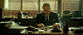 Movie still from “22 Bullets” (2010), directed by Richard Berry – A man sitting at a desk looking at papers on top of a table; Medium shot, Low angle