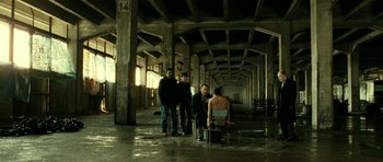 Movie still from “22 Bullets” (2010), directed by Richard Berry – A group of men standing around a chair in an empty building; Wide shot, High angle