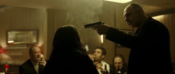 Movie still from “22 Bullets” (2010), directed by Richard Berry – A group of people in a room smoking and holding guns; Medium shot, Over the shoulder angle