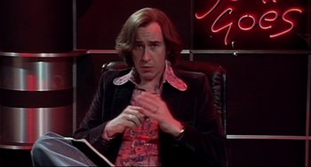 Movie still from “24 Hour Party People” (2002), directed by Tom Bruggen – A man sitting in a chair in front of a neon sign; Medium shot, High angle